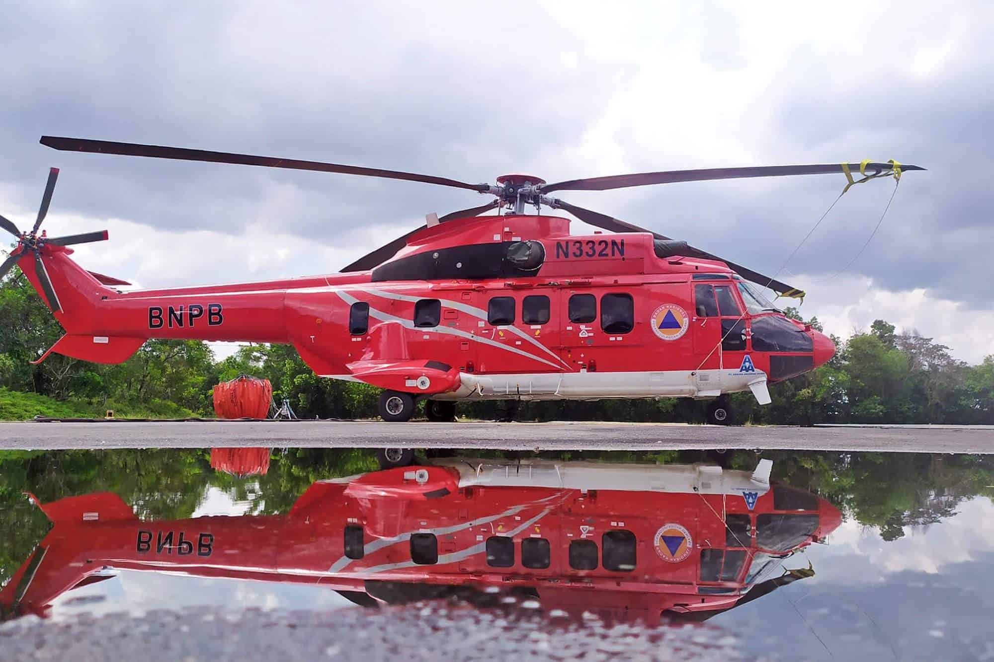 A red helicopter is parked on the ground, reflected clearly in a puddle, with trees in the background.