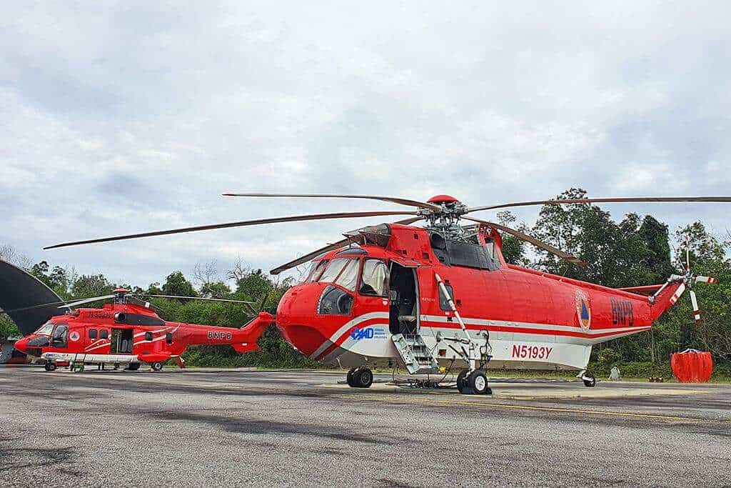 Two red rescue helicopters parked on a tarmac, with trees and cloudy sky in the background.