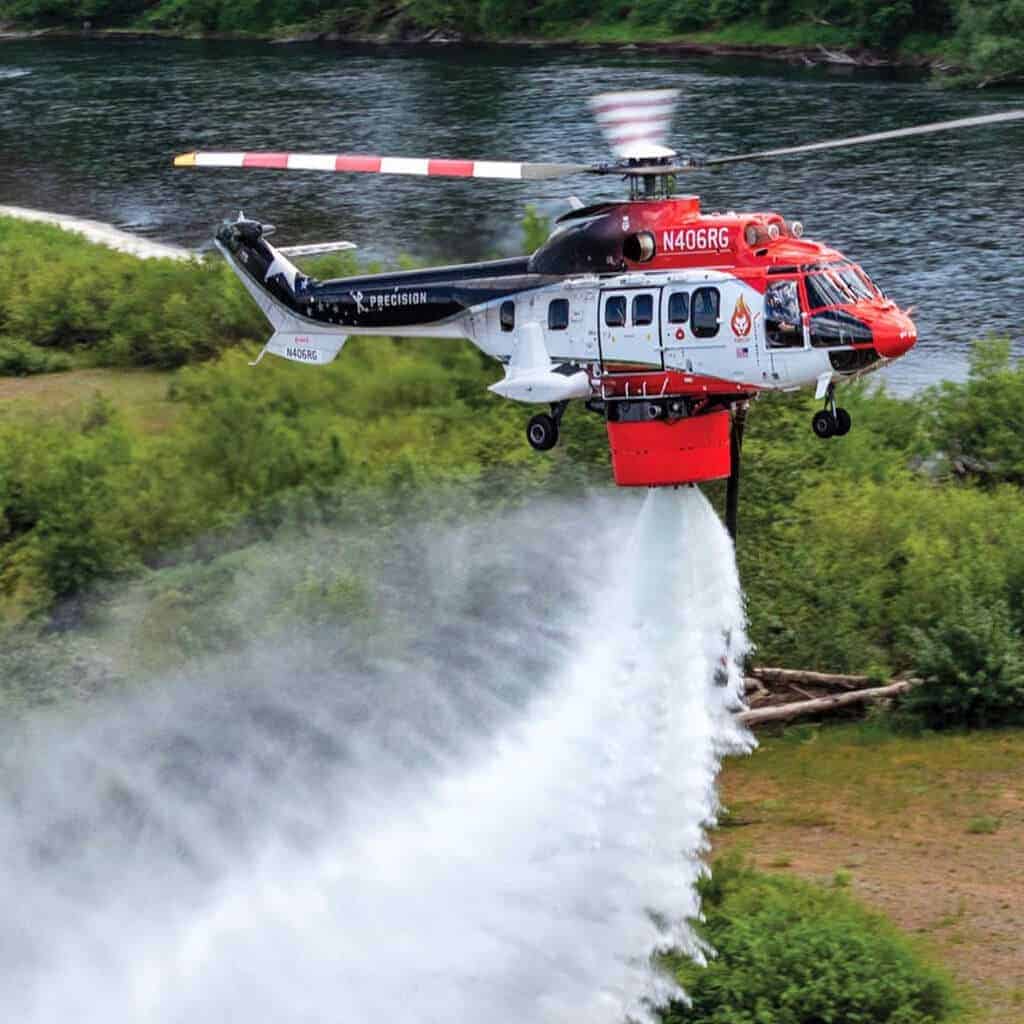 A helicopter drops water from a large bucket over a grassy area near a river.