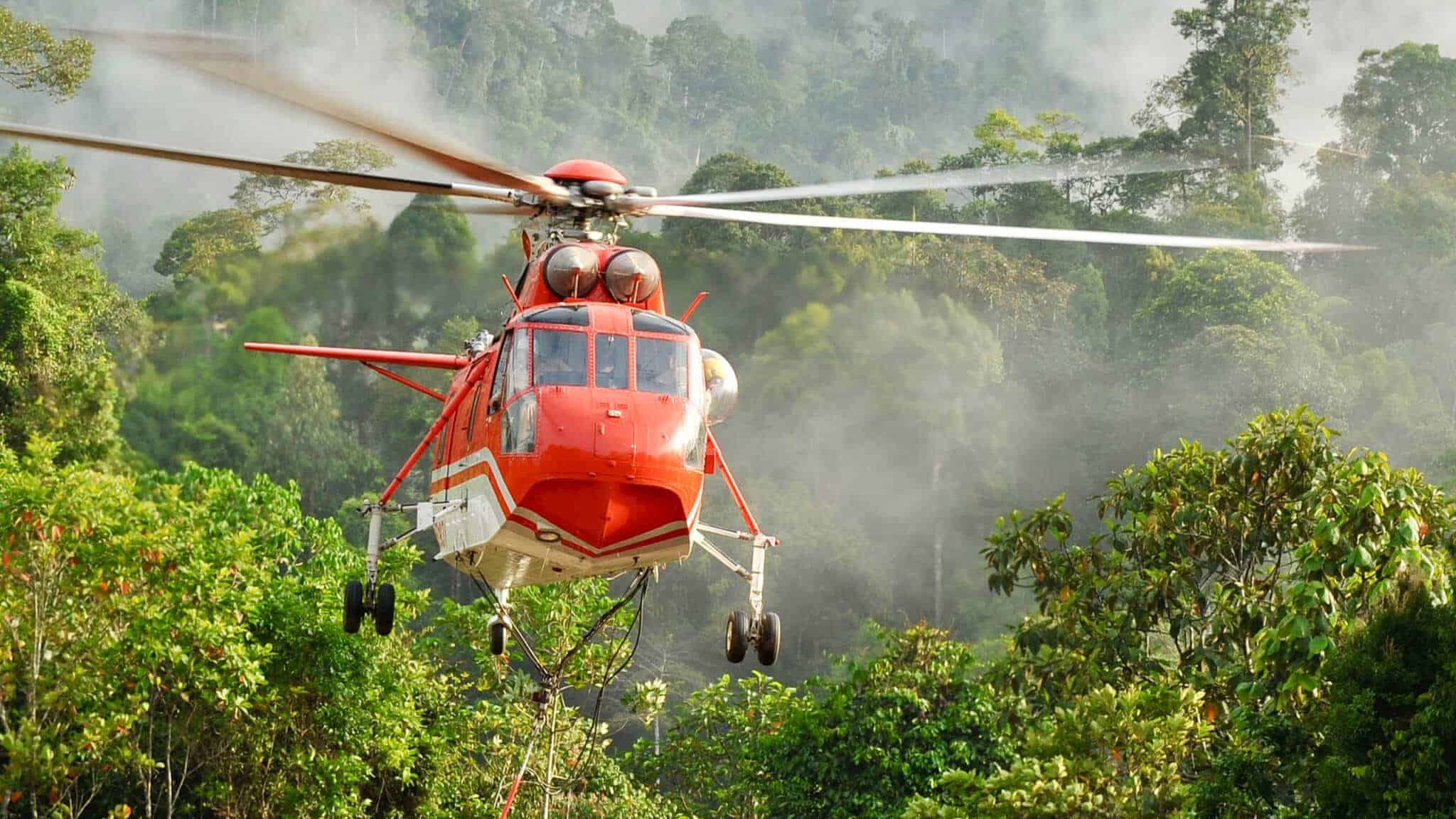 A red heavy lift helicopter from Nomad Aviation hovers above dense green jungle foliage, with trees and mist in the background—typical of South East Asia disaster relief operations.